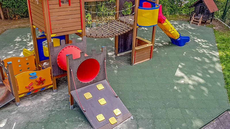 Auf grünen Fallschutzplatten mit Puzzleverzahnung steht ein kombiniertes Spielgerät mit Kletterturm, Hängebrücke und Rutsche.