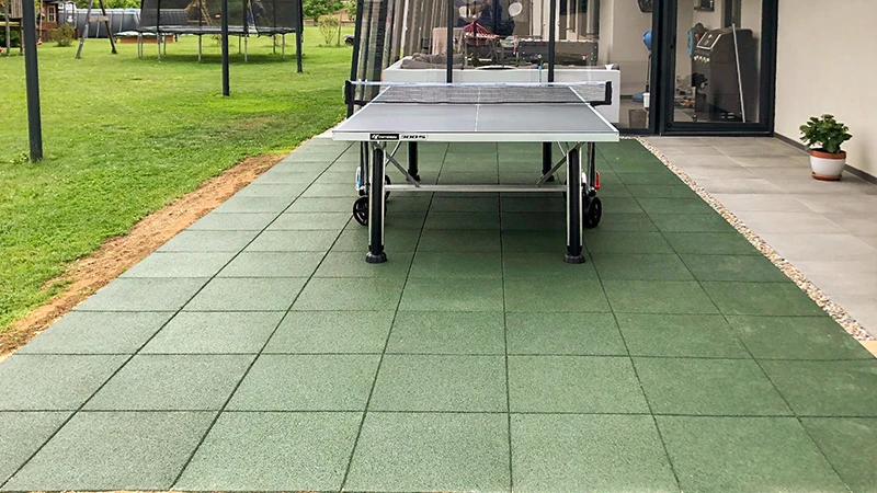 På en terrasse med grønne WARCO-fliser af PU-bundet gummigranulat er et bordtennisbord placeret foran et luksuriøst hus. I haven står en trampolin og andet legeredskab, som indbyder til aktiv afslapning og bevægelse. De robuste, skridsikre fliser giver komfort og sikkerhed. Den elastiske overflade skåner leddene, mens den vedligeholdelsesvenlige struktur bevarer et pænt udseende i al slags vejr.