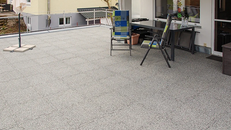 På en tagterrasse er der gråmelerede WARCO-terrassefliser af PU-bundet gummigranulat. Deres puslesamling sikrer en nøjagtig pasform og forener vejrbestandighed med lang levetid. Det formstabile materiale dæmper trinlyd og bidrager til et ryddeligt, velplejet udtryk. Samtidig giver det skridsikkerhed og høj gangkomfort, hvilket gør belægningen særligt brugervenlig.