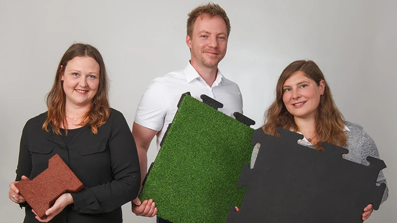 Med et venligt smil fremviser tre WARCO-konsulenter forskellige gulvfliser af PU-bundet gummigranulat i brune, grønne og sorte nuancer. Disse fliser er særligt velegnede til altaner og giver en skridsikker, slidstærk overflade. Hver variant har sit eget design og er nem at montere. Med deres ekspertise hjælper konsulenterne dig med at vælge den rette gulvbelægning for en sikker og indbydende altan. Fliserne kræver minimal vedligeholdelse og giver god komfort.