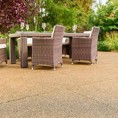 Solrig terrasse i haven med beige-brune WARCO-fliser fremstillet af PU-bundet gummigranulat. Den skridsikre, vandgennemtrængelige overflade giver en behagelig gangoplevelse og dæmper trinlyde. Et robust havemøbelsæt med flettede stole og fyldte vinglas indbyder til afslapning. Frodige buske og farverige blomster skaber en naturlig stemning. Det elastiske, frostsikre materiale bevarer sin form og er velegnet til helårsbrug.
