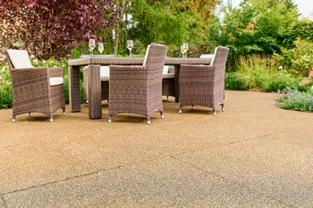 Solrig terrasse i haven med beige-brune WARCO-fliser fremstillet af PU-bundet gummigranulat. Den skridsikre, vandgennemtrængelige overflade giver en behagelig gangoplevelse og dæmper trinlyde. Et robust havemøbelsæt med flettede stole og fyldte vinglas indbyder til afslapning. Frodige buske og farverige blomster skaber en naturlig stemning. Det elastiske, frostsikre materiale bevarer sin form og er velegnet til helårsbrug.