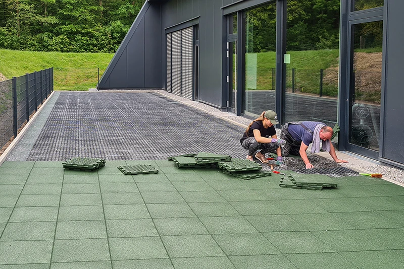 Op ad en mørk, moderne facade installerer en kvinde og en mand grønne WARCO-terrassefliser fremstillet af PU-bundet gummigranulat. Fliserne lægges tæt for at skabe en skridsikker, vejrbestandig og holdbar overflade på terrassen. Det robuste materiale giver komfort og beskyttelse i al slags vejr. Den enkle installation muliggør individuel tilpasning og giver enhver terrasse et kvalitetspræget, nutidigt udtryk. Desuden sikrer flisernes styrke langvarig funktion.