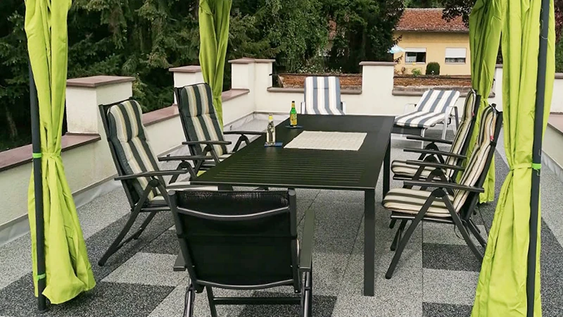På en tagterrasse ligger et gulv af WARCO-terrassefliser i lys- og mørkegrå marmorering, der minder om granit. Et baldakin dækker siddeområdet på det elastiske, vejrbestandige underlag. De skridsikre, stødabsorberende fliser kan lægges uden fast klæbning og dæmper trinlyd. Resultatet er en vedligeholdelsesnem overflade, der egner sig perfekt til afslappende timer under åben himmel.