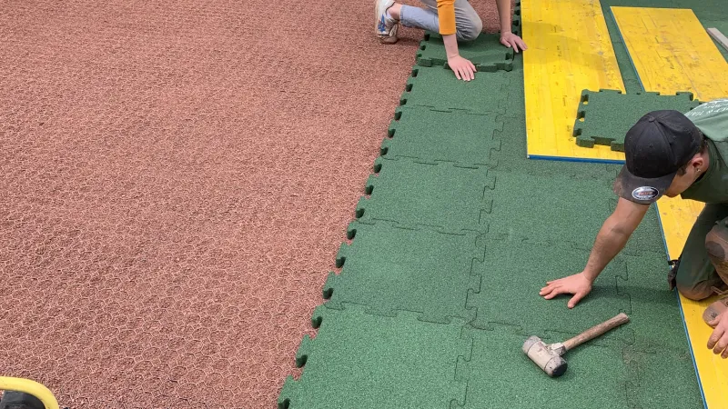 Ein Mann verlegt grüne Fallschutzplatten mit Puzzleverzahnung, d.h. in die Seiten eingeformtem Schwalbenschwanz-Verbindungssystem. Die Fallschutzplatten werden auf einer Tragschicht aus Kunststoffwabengitter verlegt. Die planeben verlegten Wabengitter sind mit rotem Splitt gefüllt.